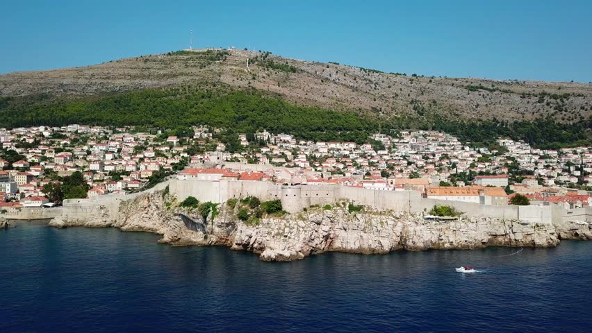 A smooth drone tracking shot moving along the majestic sea walls of Dubrovnik, Croatia, showing the fortifications on cliffs with a mountain backdrop.
