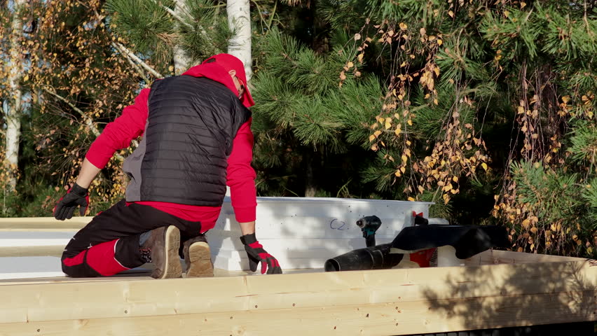 A person is building something in a forest area using wood and tools. The person wears a red jacket and works carefully.