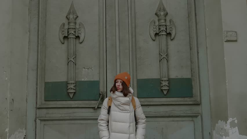 A young woman stands in front of a massive metal door decorated with axe motifs, wearing a yellow hat and white jacket, creating a bold visual contrast.