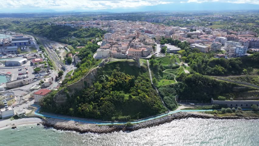 Aerial panorama of Ortona port showing turquoise sea, peaceful ripples near the shore, moored boats, large ships and detailed harbor facilities under a bright sky with soft clouds creating a serene co
