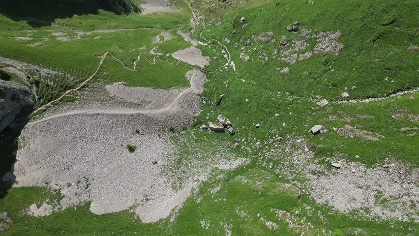 Smooth drone pullback over winding alpine hiking trails revealing a hazy valley with patchwork farmland near Saxer Lucke in the Swiss Alps.