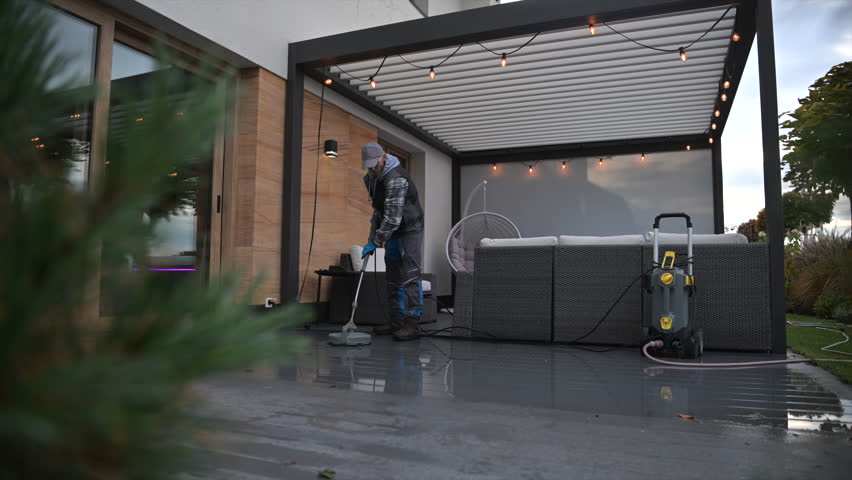 A person is using a pressure washer to clean a patio area outside a house. The patio is tidy and has furniture.