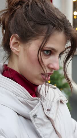 Charming young woman with her hair in a bun playfully takes off her winter puffer jacket, smiling and posing for the camera on a city street decorated for Christmas holidays