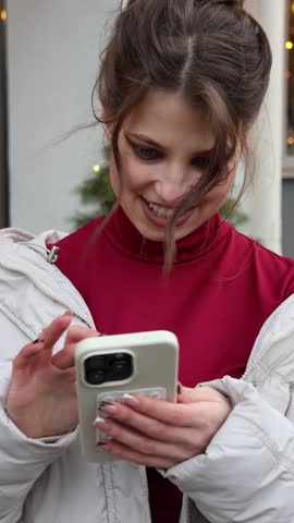 Portrait of a happy young woman using a smartphone on a city street. A beautiful girl in a winter jacket is texting on her phone with a smile, with Christmas decorations in the background.