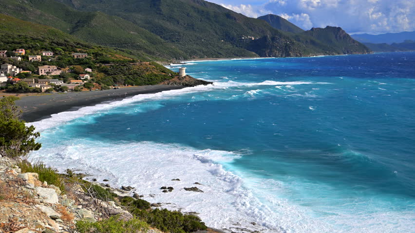 Sunlight shines on waves crashing against the black sand beach in Corsica with hills in the background.