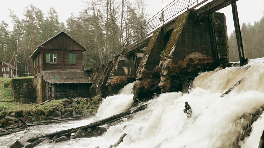 View of an old hydroelectric power station with a rushing river, historic dam, and a small wooden house nearby during overcast weather.