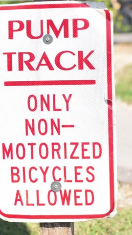 Verti al screen closeup pump track sign Johnson City park, red and white metal notice stating only non-motorized bicycles allowed, weathered bolt and post, grassy background, daytime