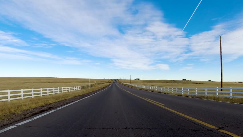 Reverse view driving down a desolate highway with blue skies some wispy clouds a jet flying overhead. Passing telephone poles and a white fence.