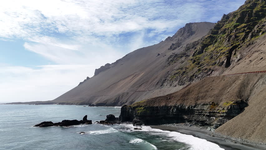 Aerial view of ocean waves hitting the rugged cliffside mountains of Iceland, revealing a winding coastal road, steep rocky slopes, and bright blue water stretching along the dramatic shoreline.