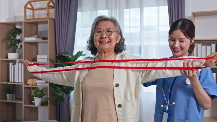 Asian Elderly woman guided by caregiver performing resistance band shoulder exercise in bright living room smiling and engaged for home physical therapy routine