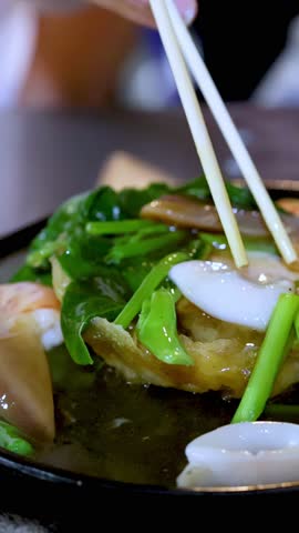 Close-up of chopsticks lifting squid from hot noodle seafood dish on sizzling plate, shallow focus