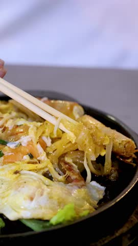 Chopsticks lift steaming Thai noodles and seafood from a sizzling hot plate under soft lighting