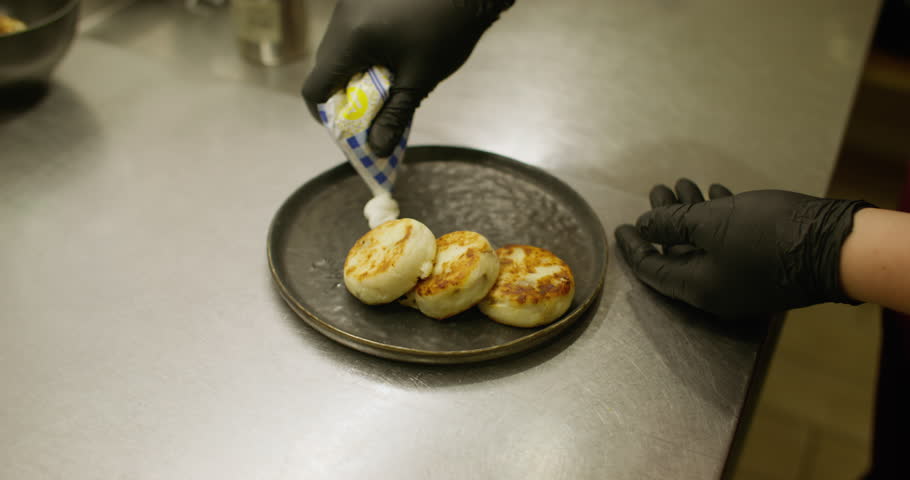 Cooking breakfast. Sweet dish. Restaurant chef adding sour cream jam to portioned cheesecakes with powdered sugar. Hands serving syrniki on plate.