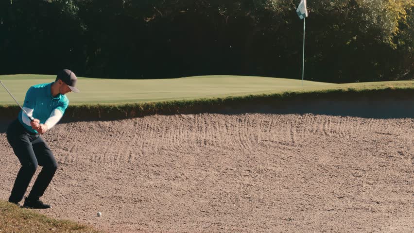 Slow-motion strike from a sand trap sends the ball onto the green beside the flag, capturing clean contact and rising sand. Male golfer.