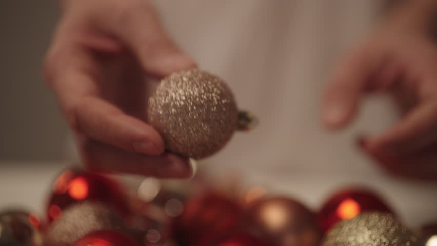 Close-up of hands holding sparkling gold glitter Christmas ornament while selecting festive decorations for holiday season, cozy warm bokeh background, preparing tree and celebrations, slow motion.