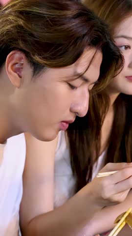 Smiling Asian couple share chopstick noodles at vibrant night market, close-up handheld evening lighting