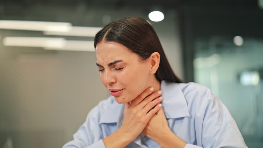 Feeling unwell, young woman suffers from a painful sore throat and persistent coughing at office desk. Depicts discomfort, illness, and health concerns, showing difficult moment.