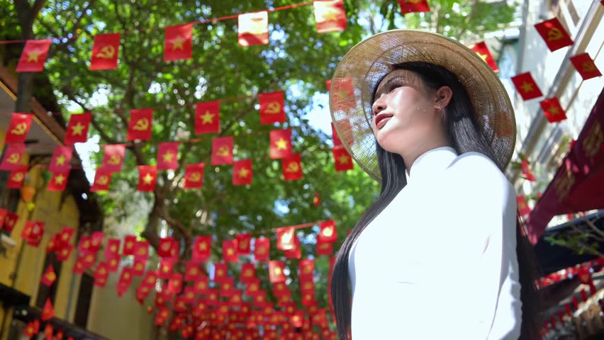 A young Vietnamese woman wearing a traditional white ao dai and a conical hat smiles gracefully under bright sunlight, in front of vibrant red Vietnam flags decorations. Travel and Vietnamese culture