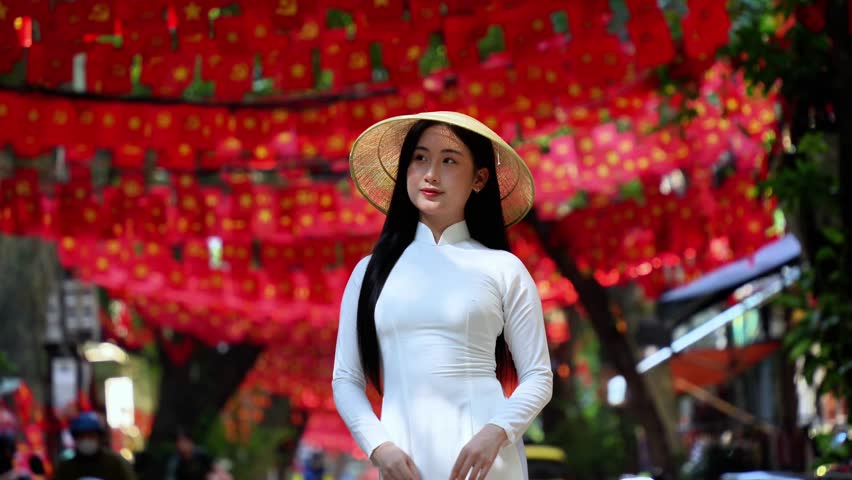 A young Vietnamese woman wearing a traditional white ao dai and a conical hat smiles gracefully under bright sunlight, in front of vibrant red Vietnam flags decorations. Travel and Vietnamese culture