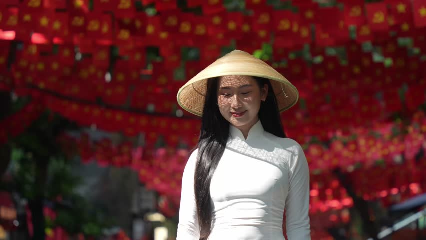 A young Vietnamese woman wearing a traditional white ao dai and a conical hat smiles gracefully under bright sunlight, in front of vibrant red Vietnam flags decorations. Travel and Vietnamese culture