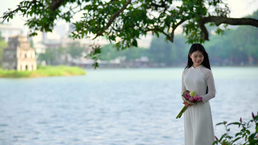 A young Vietnamese woman in a white ao dai standing by Hoan Kiem Lake, holding a bouquet of pink lotus flowers. Travel and Vietnamese culture