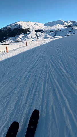 Pov footage of a person skiing down a perfectly groomed slope at a mountain resort on a sunny day, with the majestic snow covered peaks of the alps visible against a clear blue sky