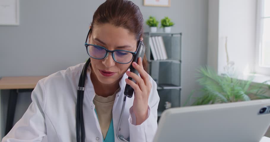 Focused young female doctor in lab coat speaking on mobile phone while checking information on laptop in clinic. Professional woman physician providing remote consultation or distant patient support.