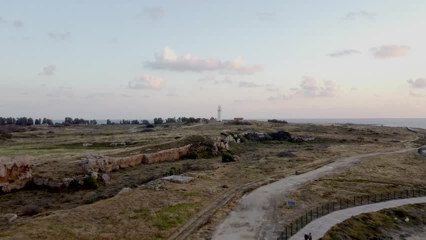 A beautiful coastal landscape showcases historic ruins and a lighthouse during sunset. Soft colors fill the sky as waves gently lap the shore, creating a peaceful atmosphere.