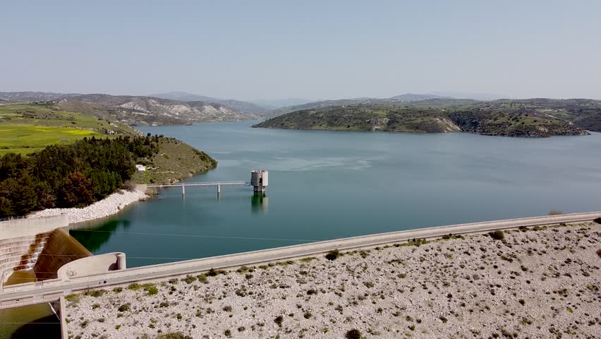 Witness the breathtaking landscape around the lake featuring a dam, lush greenery, and rolling hills. The peaceful water reflects the clear sky and distant mountains.
