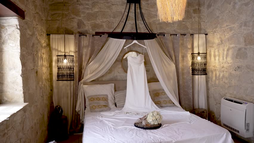 A charming bedroom features a stunning wedding dress hanging above a neatly made bed. The cozy atmosphere is enhanced by warm lighting and natural stone walls.
