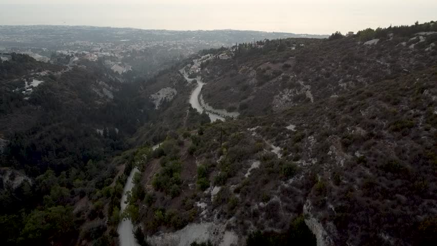 Enjoy a breathtaking aerial view of a winding road surrounded by hills in Cyprus, capturing the serene beauty of nature as the sun sets over the landscape.