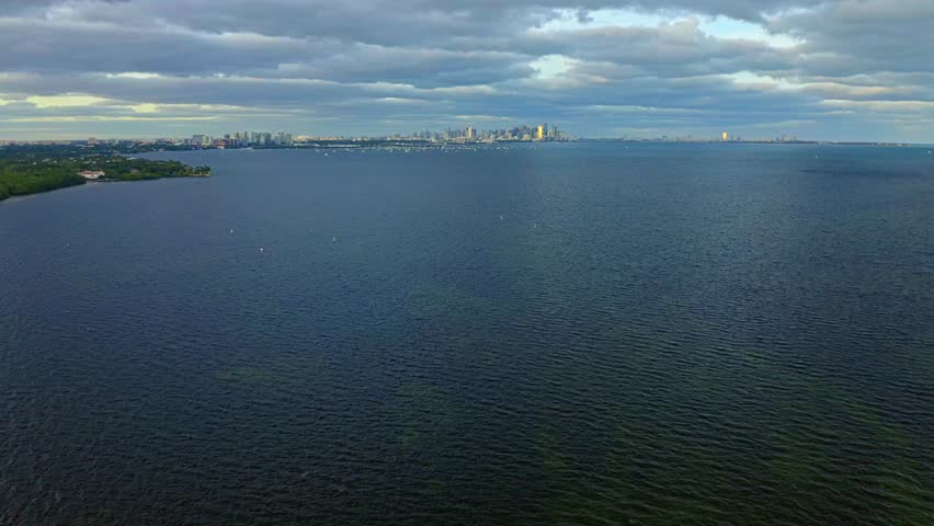 Calm Biscayne Bay stretches toward the distant Miami skyline beneath layered evening clouds, creating a wide coastal panorama from the Matheson Hammock Park shoreline.