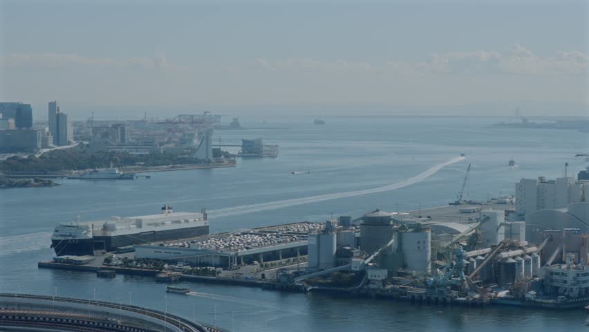 High-angle view over Tokyo Bay showing ships moving across calm water, industrial waterfront structures, and distant city skyline.