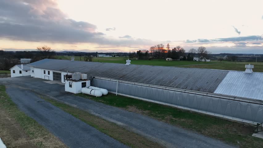 Aerial view of rural America at sunset, with a long agricultural barn, open farmland, scattered homes and wide green fields under soft evening clouds. Pennsylvania USA in Autumn.