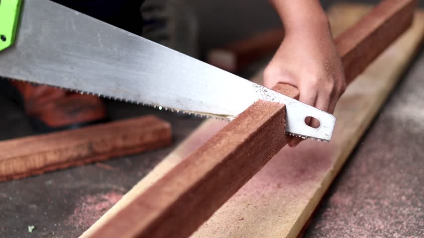 Close-up of a sharp hand saw blade cutting through wood, capturing sawdust flying in the air against a dark, blurred background.