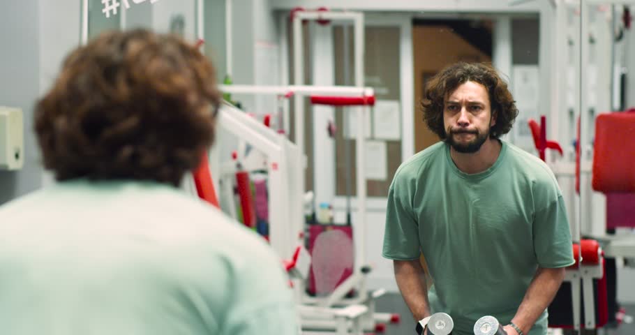 Slow motion 4k footage of a Caucasian man during gym workout in front of a mirror. Standing dumbbell raises for deltoids. Deltoid workout with dumbbells. Zoom in video.