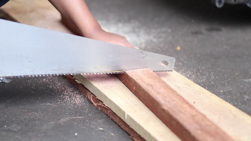 Close-up of a sharp hand saw blade cutting through wood, capturing sawdust flying in the air against a dark, blurred background.