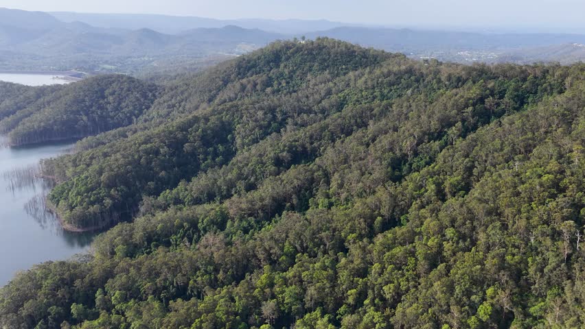 Drone glides above dense Australian bushland, revealing forested hills, lake, and distant mountains