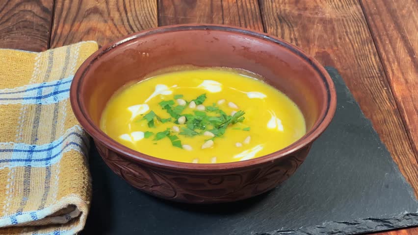 Adding diced croutons to the pumpkin soup puree with red lentil, seasoned with pine nuts, sour cream and fresh chopped greens in clay bowl on the rustic table, view close-up
