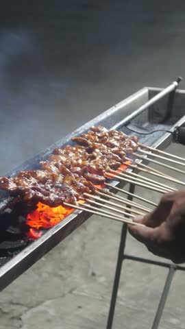 The process of grilling chicken satay over hot coals on a long grill. Rows of skewers are golden brown with a glistening of spices, while flames and smoke rise from the grill.