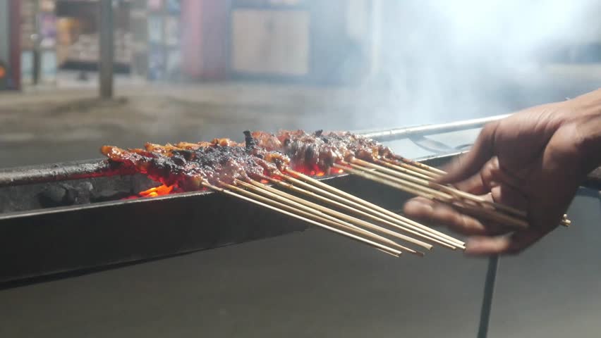 The process of grilling chicken satay over hot coals on a long grill. Rows of skewers are golden brown with a glistening of spices, while flames and smoke rise from the grill.