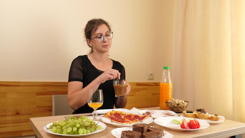 A young woman in a black T-shirt and glasses is drinking coffee and eating pastries while sitting at a table.