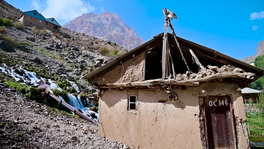 A view of a small shack on a hillside above a clear mountain stream in the Seven Lakes region of the Fann Mountains near Panjakent, Tajikistan, with rocky terrain and sparse vegetation.