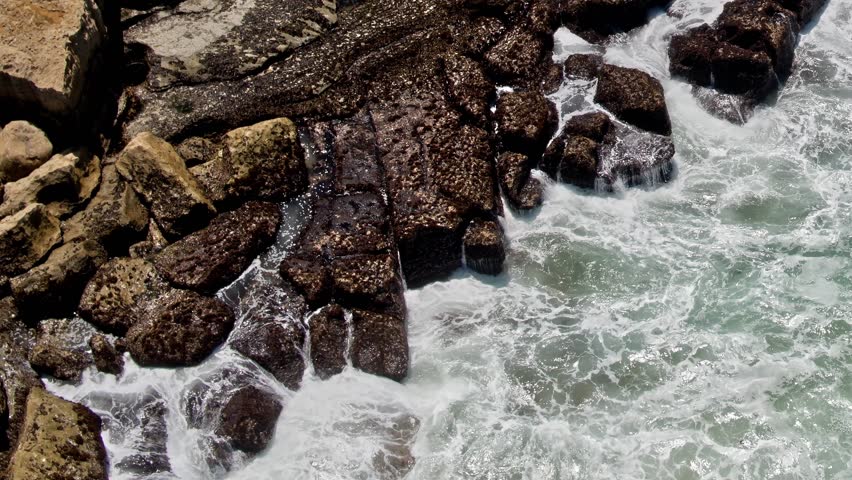 Waves roll in, crashing against rocky shores in Portugal. The water churns and moves in different patterns under the bright daytime sky.