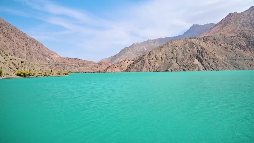 A turquoise water view of Iskanderkul Lake in the Fann Mountains of Tajikistan, showing rocky shoreline and steep alpine slopes in a remote Central Asia setting.