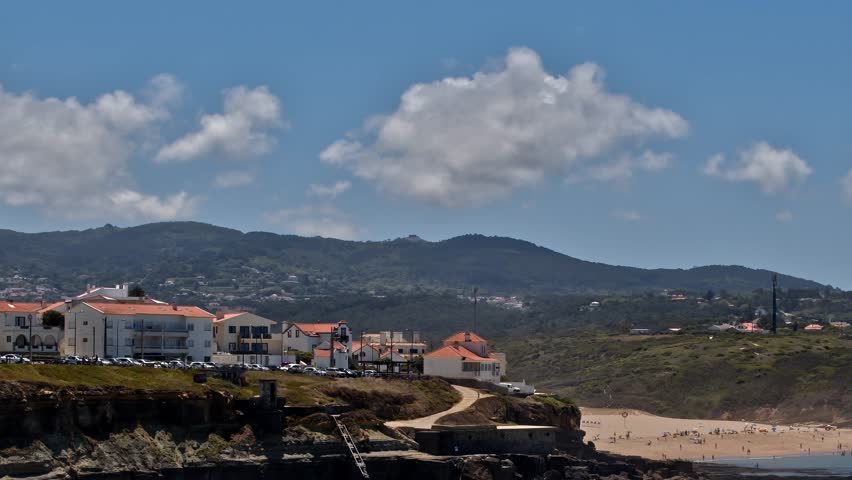 A beach in Portugal with white sand, hills in the background, and buildings along the shore. People walk on the sand under a blue sky with clouds.