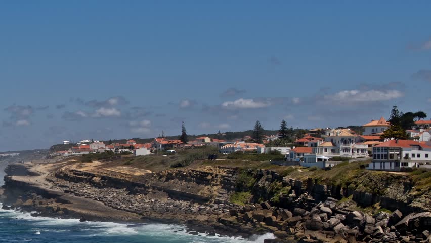 The scene shows a coastal area in Portugal with houses lined along the shoreline. Waves crash against the rocks below. The sky is clear, and the landscape is inviting.