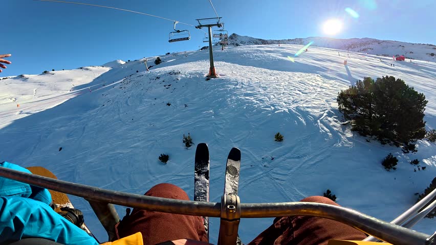 Pov footage of a skier riding a chairlift up a snow covered mountain at a busy ski resort on a bright, sunny day, with ski tracks visible on the slopes below and lens flare from the sun
