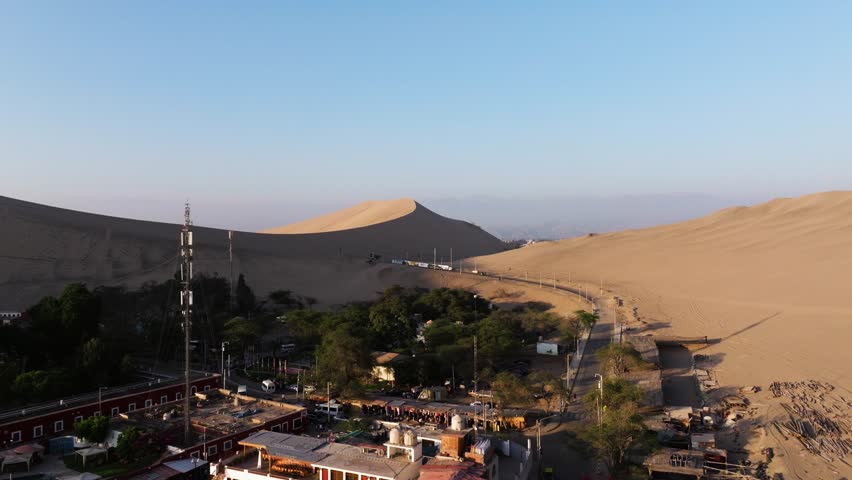 Drone ascend over desert valley revealing Huacachina oasis tucked below sweeping dunes under clear sky