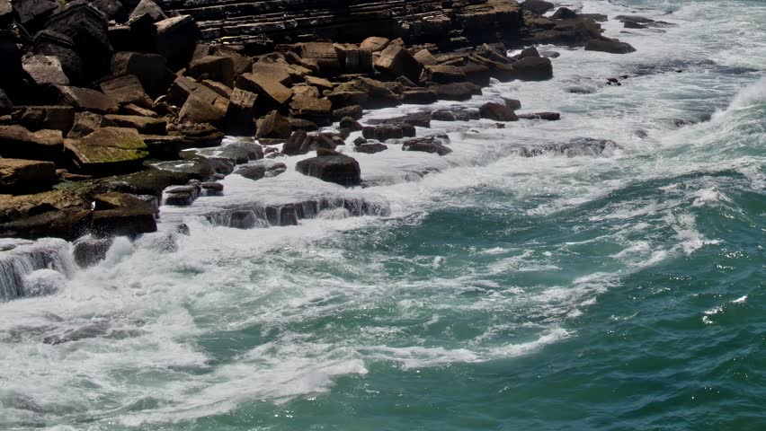 Waves crash against rocks along the coastline of Portugal. The water is blue, and the weather is clear, capturing the natural movement of the sea.